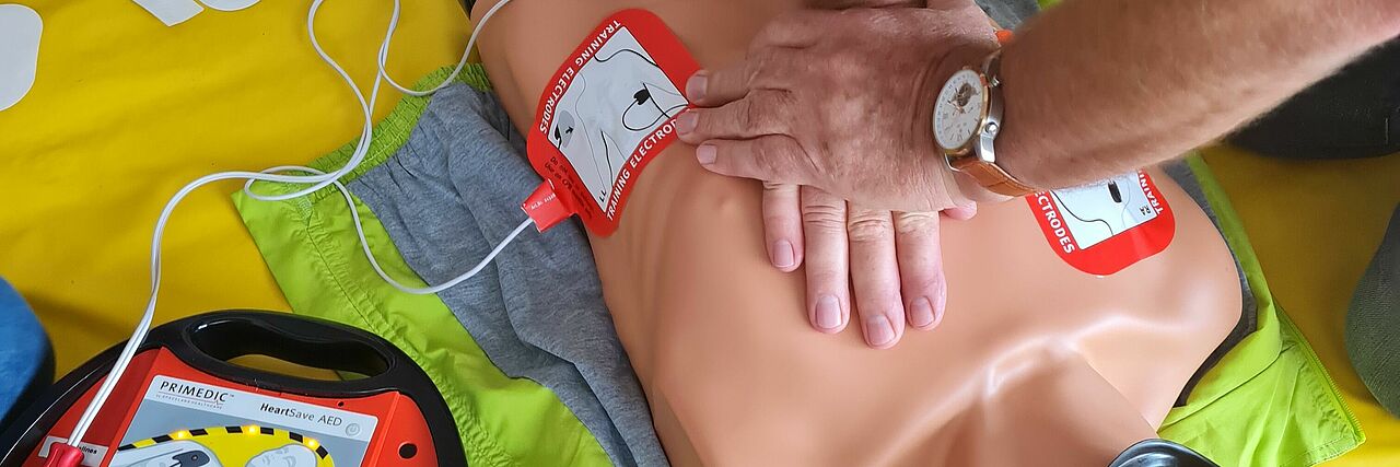 Hände sind zu sehen, wie sie an einer Wiederbelebungspuppe Herz-Druckmassage üben, daneben ist ein Defibrillator zu sehen
