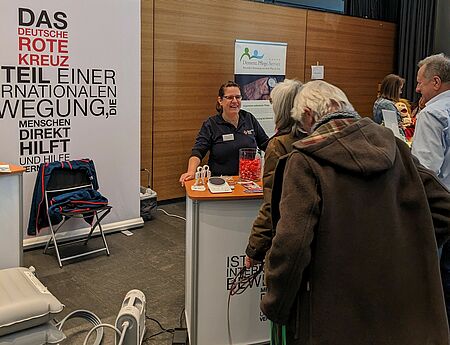 Eine Rotkreuz-Mitarbeiterin steht an einem Infostand und erläutert mehreren Menschen unsere Angebote