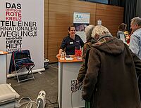 Eine Rotkreuz-Mitarbeiterin steht an einem Infostand und erläutert mehreren Menschen unsere Angebote