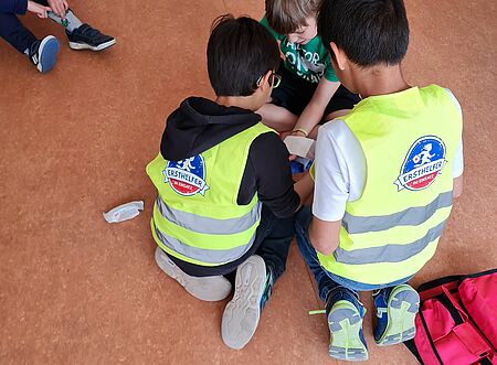 Zwei Kinder in Warnwesten helfen einem verletzten Kind