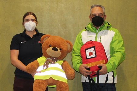 Schularbeitsreferentin Anna Pegios und ein Vertreter der Barmer stehen mit einem Teddy mit Warnweste und einem Juniorhelferrucksack vor der Kamera.