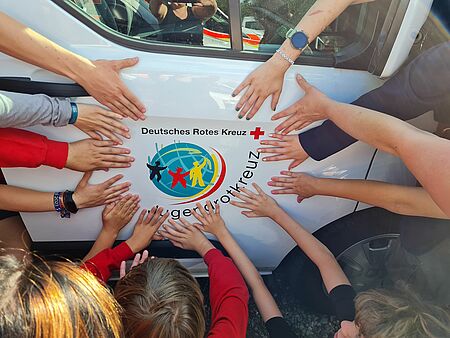 Viele Kinderhände bilden einen Kreis um das JRK-Logo