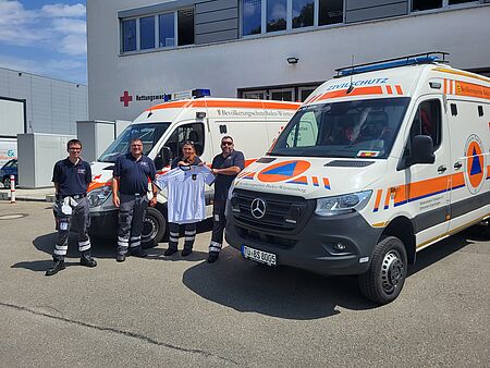 Einsatzkräfte des Sanitätsdienstes stehen mit einem Fußballtrikot vor Einsatzfahrzeugen