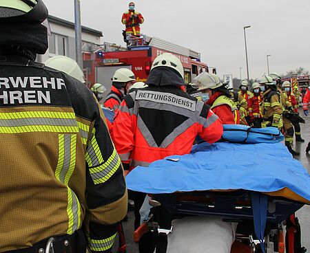 Einsatzkräfte von DRK und Feuerwehr stehen mit einer Trage am Einsatzort und warten auf die Übergabe eines Verletzten