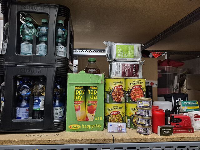 Man sieht ein Regalbrett, das mit Getränken, Lebensmitteln, Medikamenten und anderen Hilfsmitteln für eine Katastrophe ausgestattet ist.